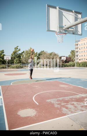 One afroamerican young man is playing basketball in a park in Madrid during summer at midday. He is shooting the ball to the basket from half distance Stock Photo