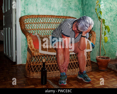A drunk senior woman with a wine bottle in her hand sitting in a sofa Stock Photo