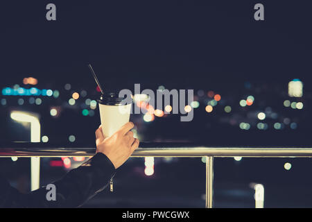 Closeup of female hand holding black craft coffee cup to take away, mock up of paper coffee cup to go outside, bokeh light. Night life style. paper Cu Stock Photo