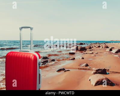 A red plastic suitcase stands on the rocky shore of a lake. Stock Photo