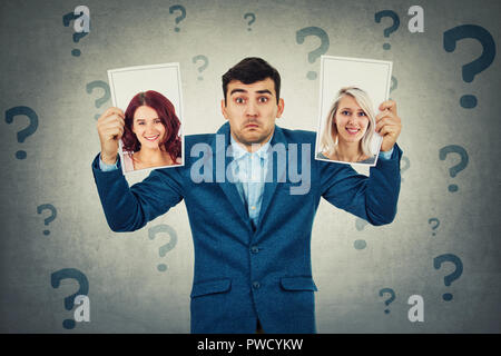 Undecided young man holding two photos has to choose between two woman, question marks around head. Girlfriend partner first date choice love and roma Stock Photo