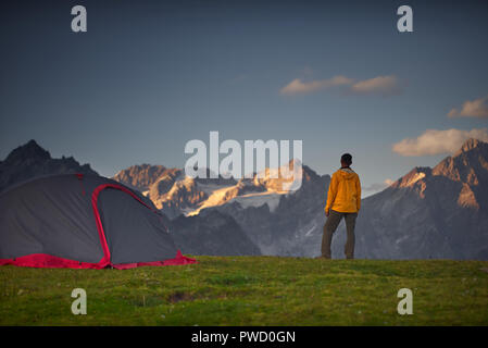 Hiker in a camp on green lawn and looking at mountains in a sumer day. Stock Photo