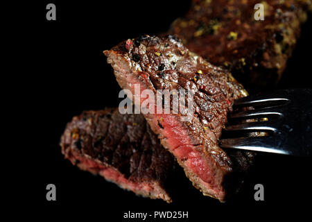 Close up of medium rare beef steak piece on a fork . Spiced with peppercorns and herbs. More fried meat on background. Isolated image with copy space. Stock Photo