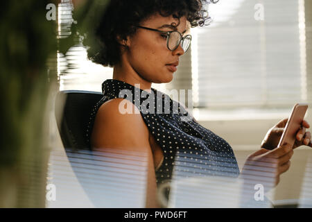Close up of young african woman using mobile phone in office. Female in casuals sitting at her desk using smartphone. Stock Photo