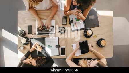 Top view of four women sitting around table in cafe with laptop, digital tablet and documents. Group of women working together in coffee shop. Stock Photo
