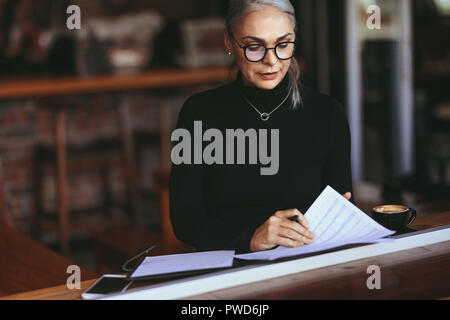 Mature woman sitting in a restaurant and doing some paperwork. Female sitting in cafe with a coffee reading some papers. Stock Photo