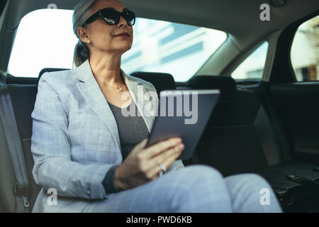 Female entrepreneur travelling to office in a luxurious car sitting on backseat with digital tablet. Senior businesswoman in car with tablet pc. Stock Photo