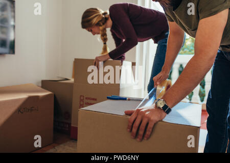 Close up of man sealing a packing box with adhesive tape while a woman placing articles in another box. Couple packing their items for moving into a n Stock Photo