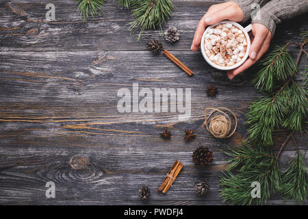 Woman's hand holding the cup of hot cocoa with marshmallows on wooden background with pine branches, pine cones, cinnamon sticks and anise stars. Traditional winter hot drink. Stock Photo