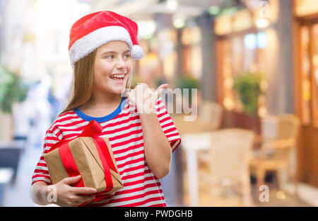 Young beautiful girl wearing christmas hat and holding gift over isolated background pointing and showing with thumb up to the side with happy face sm Stock Photo