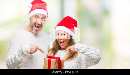 Middle age hispanic couple wearing christmas hat and holding gift over isolated background very happy pointing with hand and finger Stock Photo