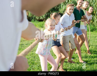 Smiling kids with moms and dads playing tug of war during joint outdoors games on sunny day Stock Photo