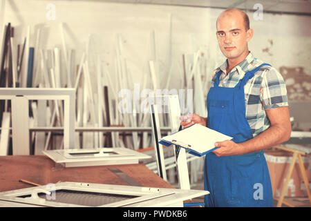 Diligent  positive focused workman in overalls controlling quality of assembly of plastic window in workshop Stock Photo