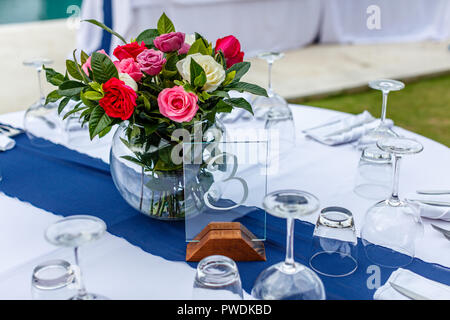 Glass round vase with bright bouquet of colorful roses on a table set up for dinner. Glass table number three. Concept of a tropical wedding. Stock Photo