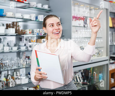 Young woman buyer holding catalogue, looking new tableware for kitchen in shop Stock Photo
