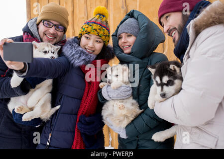 Group of young people playing with adorable husky puppies and taking selfies while enjoying nice winter day outdoors Stock Photo