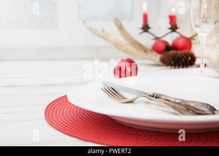 Christmas place setting with white dishware, cutlery, silverware and red decorations on wooden board. Christmas candlestick with red candles as decor. Stock Photo