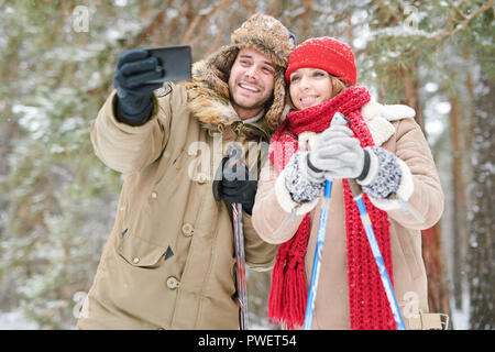 Waist up portrait of active young couple taking selfie photo via smartphone while enjoying skiing in snowy winter forest during date Stock Photo