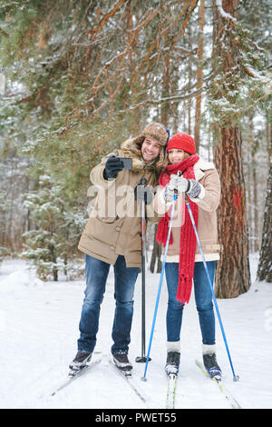 Full length portrait of active young couple taking selfie photo via smartphone while enjoying skiing in snowy winter forest during date Stock Photo