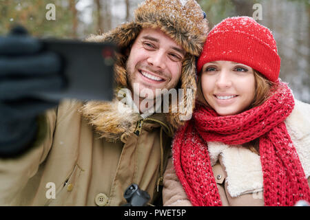 Portrait of happy young couple taking selfie photo via smartphone while enjoying date in winter forest Stock Photo