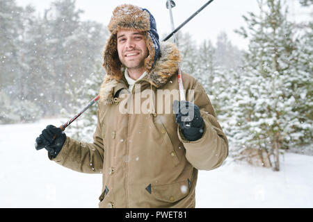 Waist up portrait of modern young man wearing fur hat smiling happily at camera while enjoying skiing in snowy winter park, copy space Stock Photo