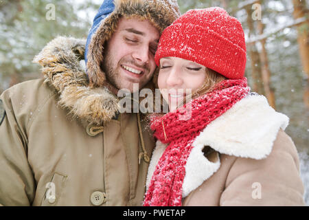 Head and shoulders portrait of loving young couple embracing tenderly and smiling with eyes closed while enjoying walk in snowy winter forest during d Stock Photo