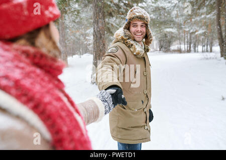 Waist up portrait of handsome young man looking at girlfriend and smiling happily while leading her through beautiful winter park, copy space Stock Photo