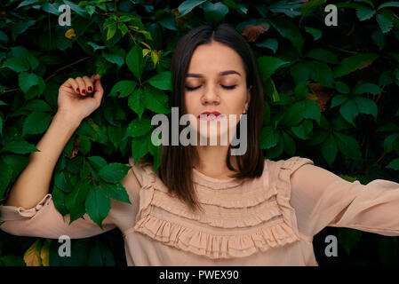 Young woman is leaning on a stone fence covered with green leaves with closed eyes. Stock Photo