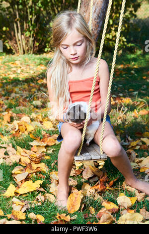 Cute child girl sitting on swing in garden and playing with guinea pig pet animal Stock Photo