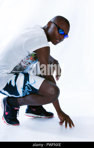 Young african male in a squatting position wearing sports clothes, sport shoes and sunglasses looking at the camera Stock Photo