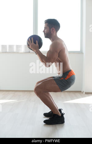 Handsome Young Man Doing Squad With Medicine Ball As Part Of Bodybuilding Training Stock Photo