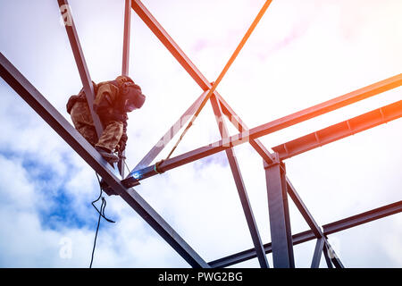 A young  man welder in brown uniform, welding mask and welders leathers, weld  metal  with a arc welding machine at the construction site, blue sparks Stock Photo