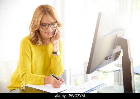 Shot of middle aged businesswoman using her cell phone and talking with client and making notes while sittin at office desk. Stock Photo