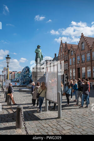 A group of young female tourists listen to a tour guide in Jan Van Eyck Square, underneath a statue of the Flemish painter, Bruges, Flanders, Belgium Stock Photo