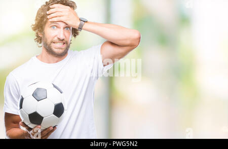 Handsome hispanic man model holding soccer football ball over isolated background stressed with hand on head, shocked with shame and surprise face, an Stock Photo