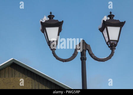 Vintage black street lamppost with two lamps and roof of building covered with snow on blue sky background Stock Photo