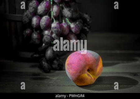 Bucket with grapes spilling from it on wooden table and peach against black background- fall harvest composition side view Stock Photo