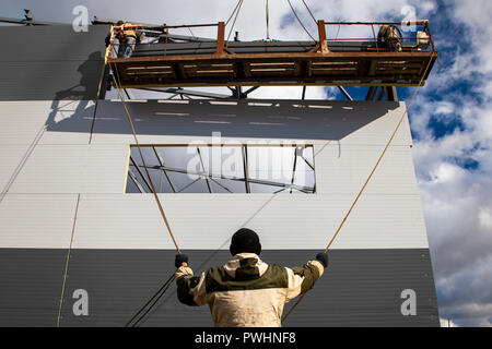 The builders are building a building of metal structures at a height in the construction cradle, in the foreground is a builder holding ropes against  Stock Photo