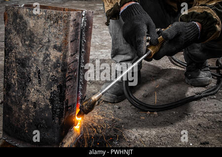 Man welder in welding mask, building uniform and black protective gloves brews metal welding machine on factory, in the background construction site Stock Photo