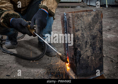 Man welder in welding mask, building uniform and black protective gloves brews metal welding machine on factory, in the background construction site Stock Photo