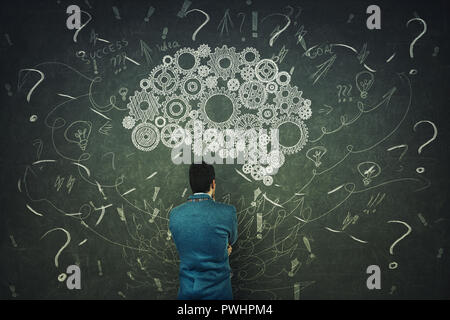 Rear view of a puzzled businessman having questions in front of a blackboard. Big chalk drawn gear brain above head, positive thinking mess as thought Stock Photo