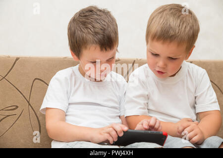 Two little boys in white T-shirts are sitting on the sofa at home. One boy shows something to another on smartphone. Stock Photo