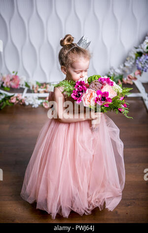 A little princess in a beautiful pink dress sniffs a bouquet of peonies, magnolia, berries and greenery against a white wall and floral arch Stock Photo