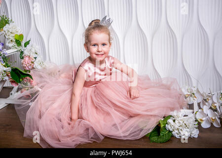 The little princess in a beautiful pink dress sits on the floor near the flower arch on a light background and smiles Stock Photo