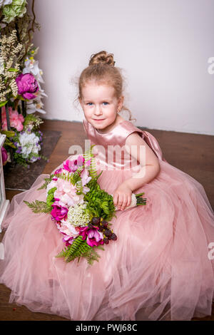 A little princess in a beautiful pink dress sits on the floor near flower stands and lanterns, holds a bouquet of peonies, magnolias, berries and greenery Stock Photo