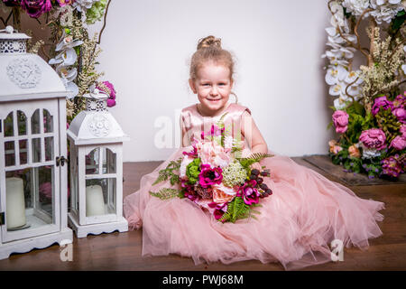 A little princess in a beautiful pink dress sits on the floor near flower stands and lanterns, holds a bouquet of peonies, magnolias, berries and greenery, and smiles Stock Photo