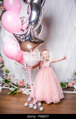 The little princess in a beautiful pink dress holds balloons, laughs and points to the side, light background Stock Photo