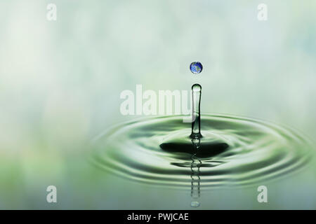 Drop of rain with blue earth image falling on smooth water surface. Green blurred pattern in background. Stock Photo