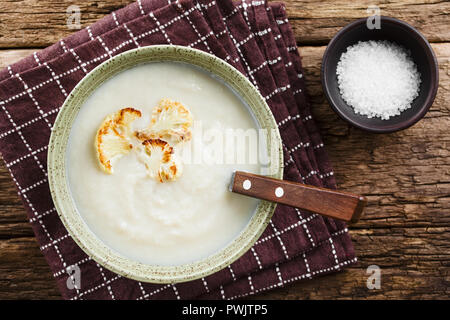 Fresh homemade cream of cauliflower soup garnished with roasted cauliflower floret slices, served in bowl with spoon, salt on the side Stock Photo