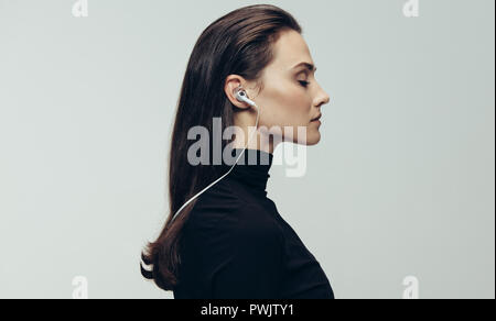Side view of woman in black dress wearing earphones with eyes closed. Woman wearing earphones against grey background. Stock Photo